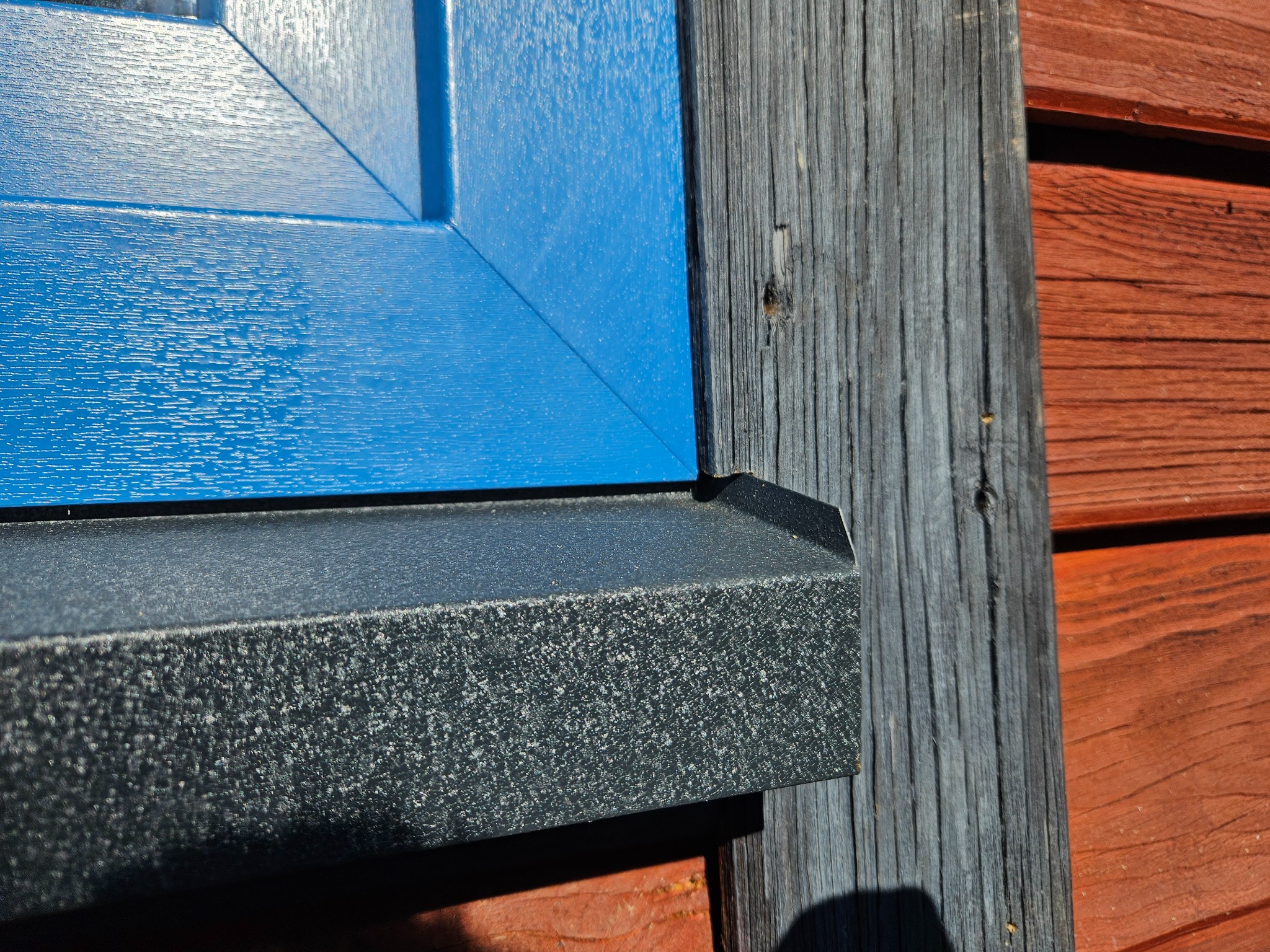 Fragment błękitnego okna z PCV, widoczny narożnik i parapet zewnętrzny w kolorze grafitowym, na tle drewnianej elewacji pomalowanej na czerwono-brązowy kolor oraz szarej deski. Widoczna faktura...
