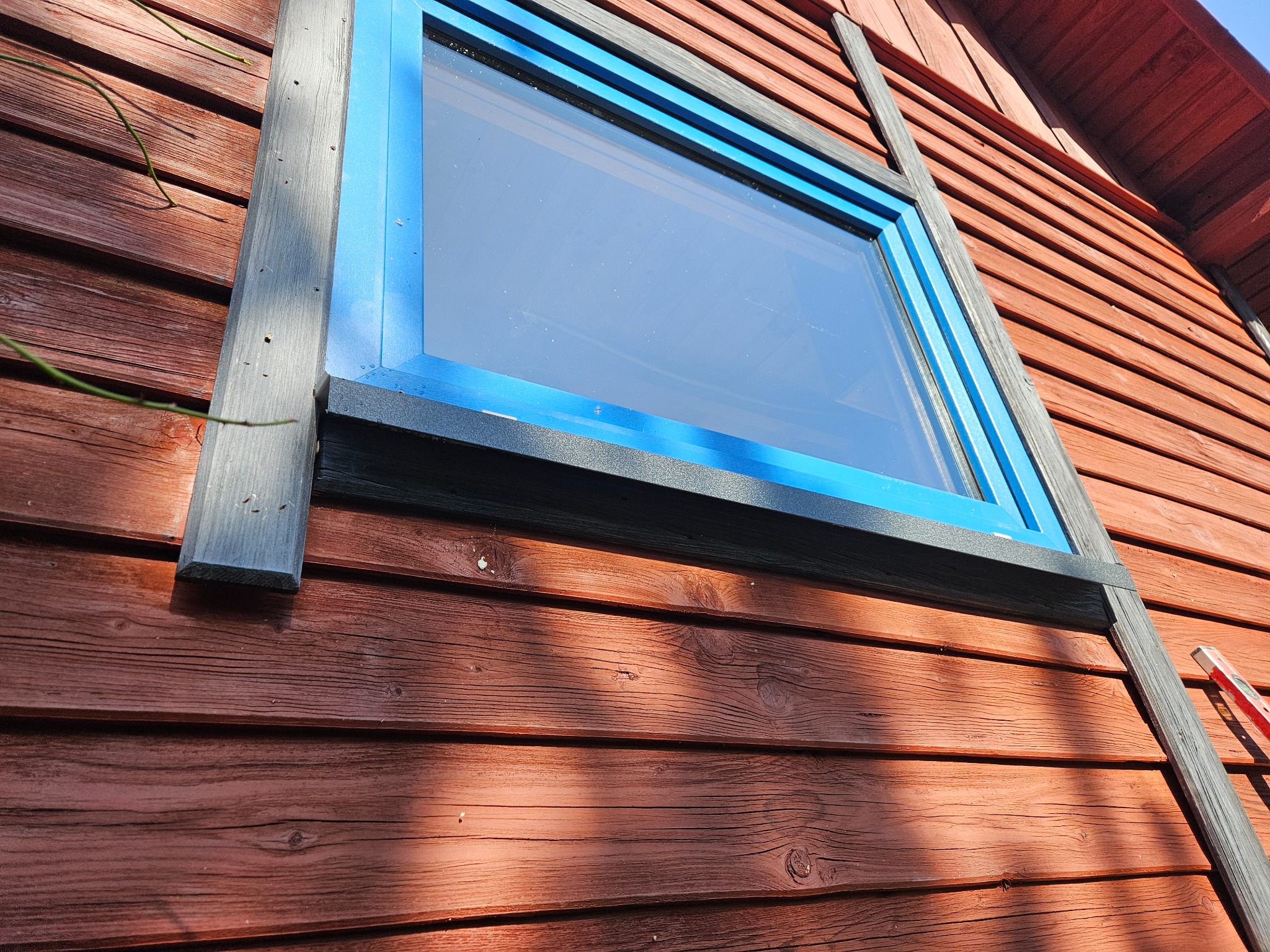 Nowe okno z niebieską ramą zamontowane w drewnianej ścianie budynku, widoczna poziomica oparta o ścianę.