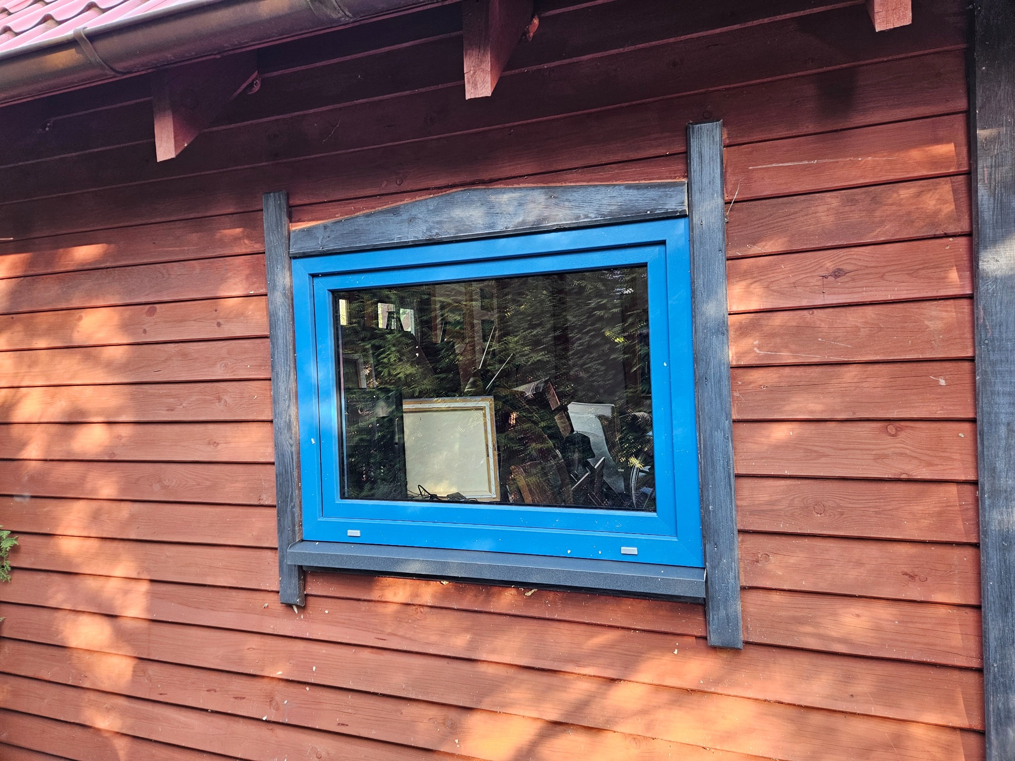 Błękitne okno PCV w drewnianej elewacji domu z widocznymi elementami ramy okiennej z czarnego drewna.