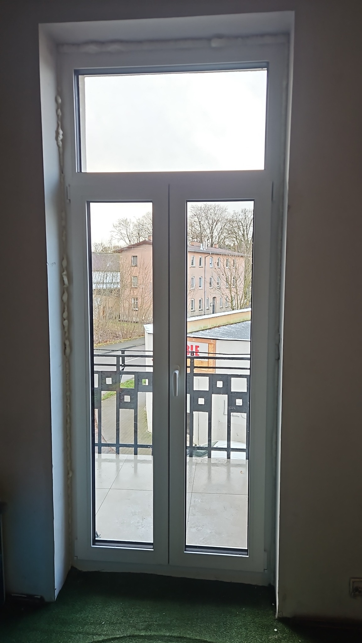 Białe, dwuskrzydłowe drzwi balkonowe PCV z widokiem na balkon i budynki w tle. Widoczne ślady pianki montażowej przy ościeżnicy. Ujęcie z wnętrza pomieszczenia.