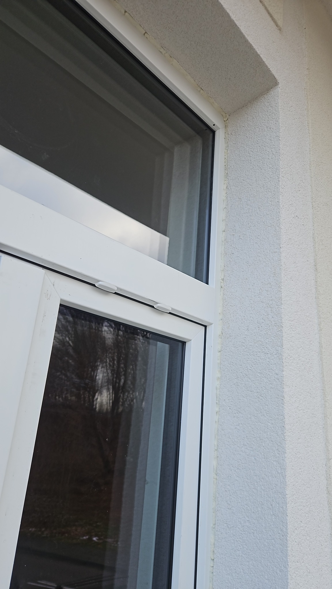Fragment białego okna PCV z widocznym uszczelnieniem silikonowym na tle jasnej, teksturowanej elewacji budynku. Widok z bliska, z boku.
