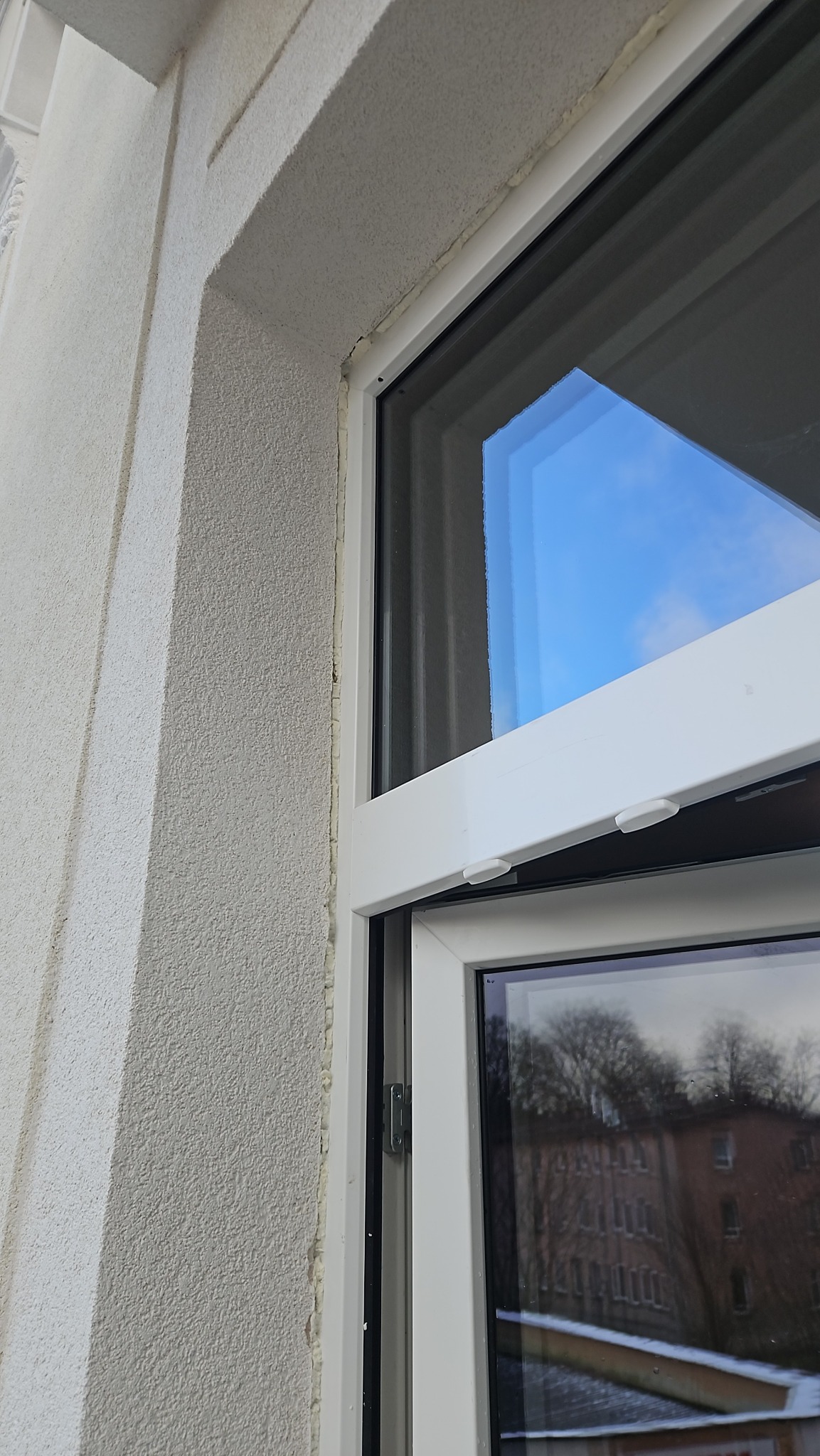 Fragment białego okna PCV z widoczną pianką montażową na styku z elewacją budynku. Widoczne odbicie nieba i drzew w szybie okiennej.