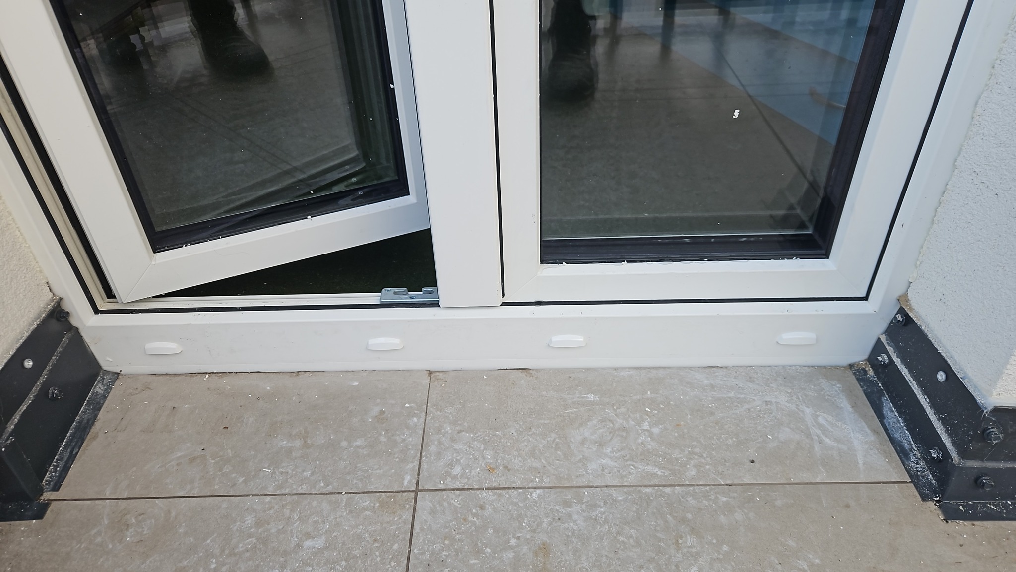 Białe okno PCV z czarnymi uszczelkami, jedno skrzydło uchylone, widoczne detale montażu i wykończenia na tle jasnej podłogi.