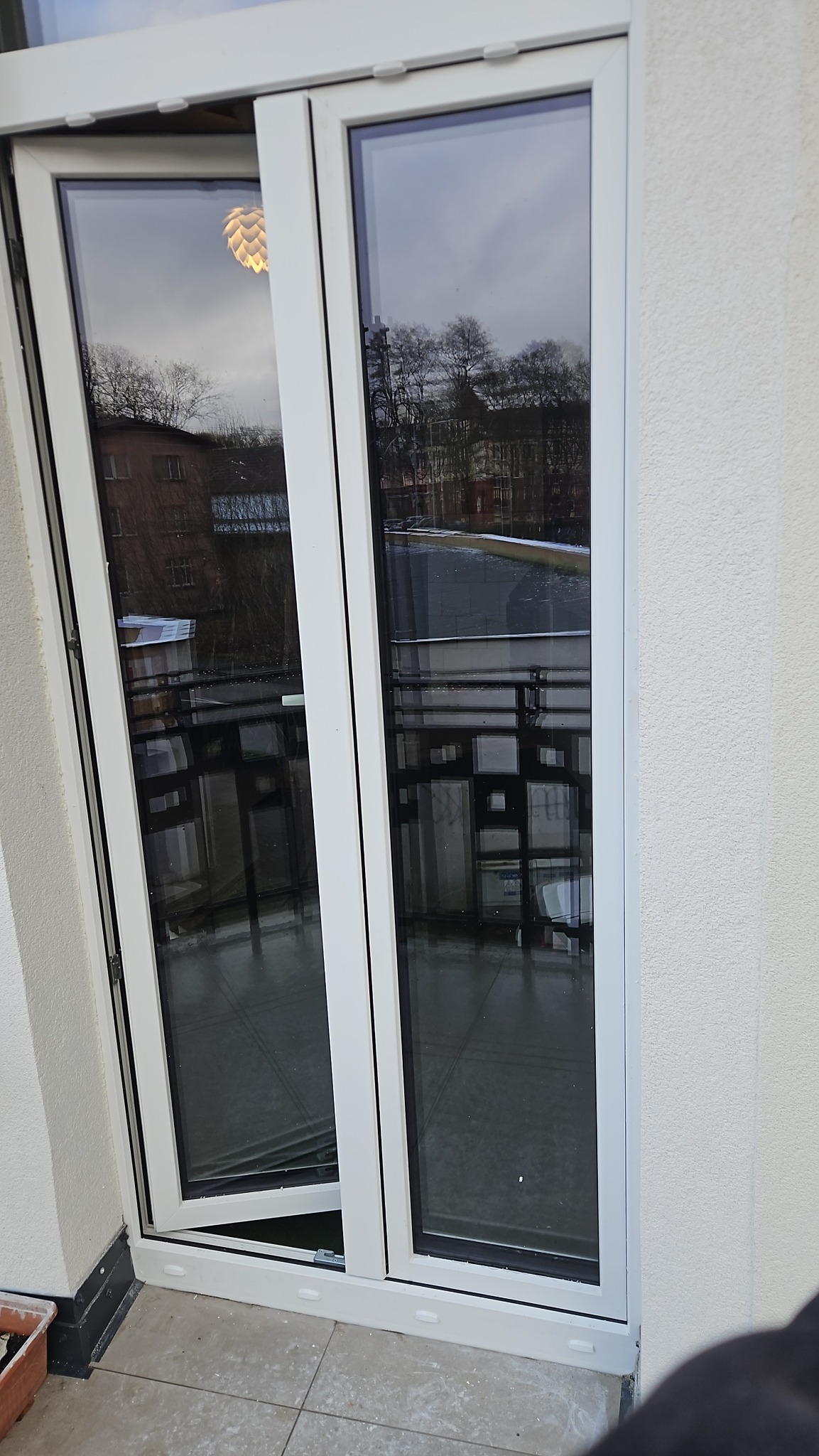 Białe, dwuskrzydłowe okno balkonowe z uchylonym jednym skrzydłem, widok na zewnątrz z odbiciem lampy w szybie. Nowoczesne wykończenie balkonu.