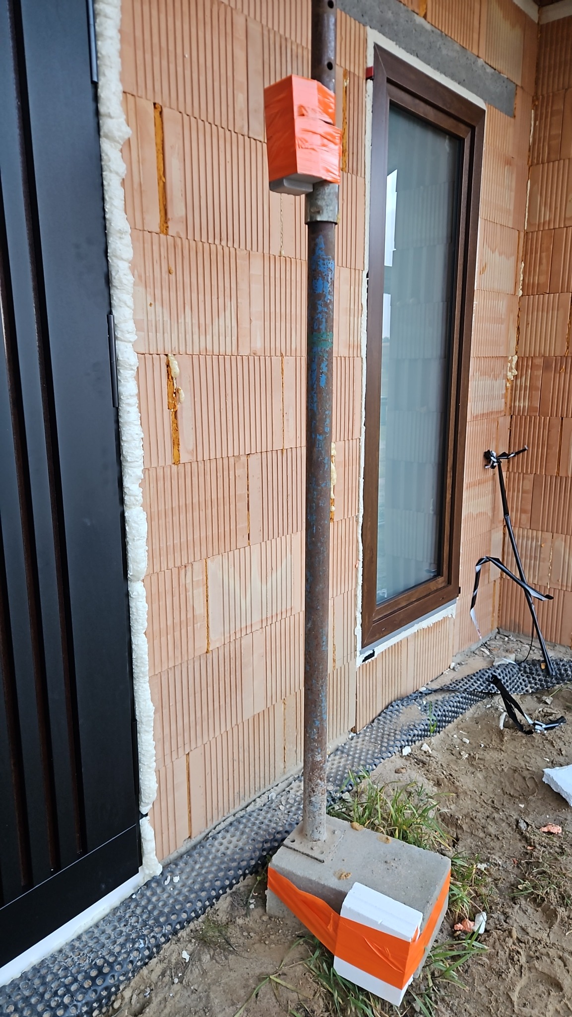 Murowana ściana z oknem i drzwiami, zabezpieczona pianką montażową. Widoczny element konstrukcyjny z pomarańczową taśmą. Etap prac wykończeniowych.