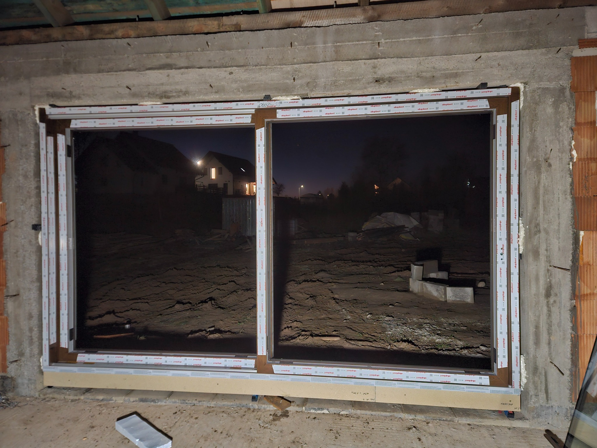 Montaż dwuskrzydłowego okna PCV w surowym murze. Widoczne profile okienne z folią ochronną, nocny widok na dom z podwórka przez szybę. Etap prac wykończeniowych.