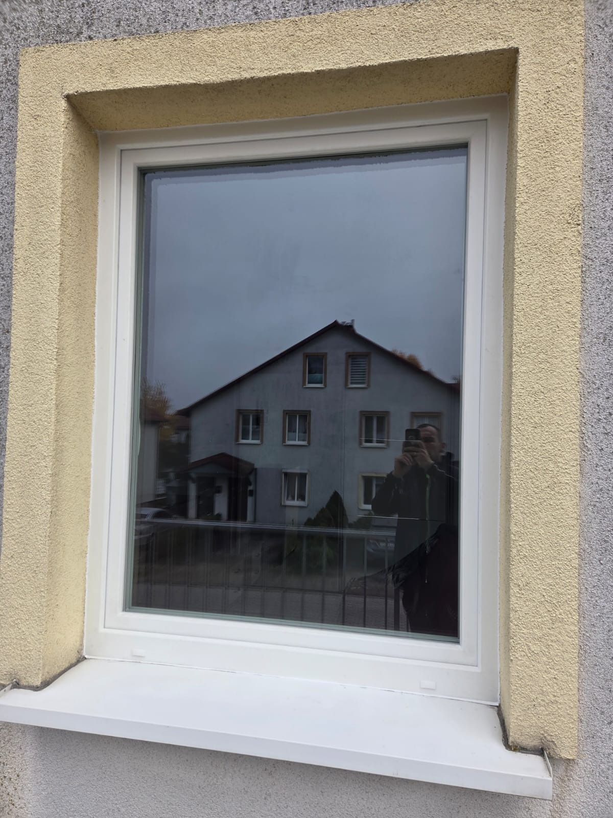 Białe okno PCV z widocznym odbiciem budynku i osoby robiącej zdjęcie. Żółta elewacja wokół okna. Nowoczesny design.