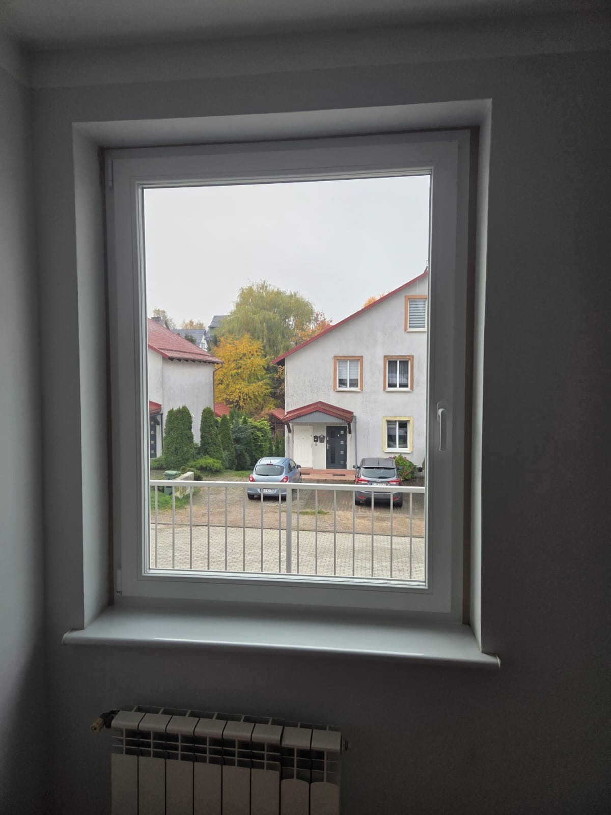 Białe okno PCV z widokiem na osiedle domów w jesiennej scenerii. Dwa samochody zaparkowane przed budynkami. Grzejnik pod parapetem.
