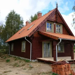 Ciemnobrązowy dom z bali z czerwonym dachem z dachówki ceramicznej, widoczny stos dachówek na ziemi, w tle brzozowy lasek.