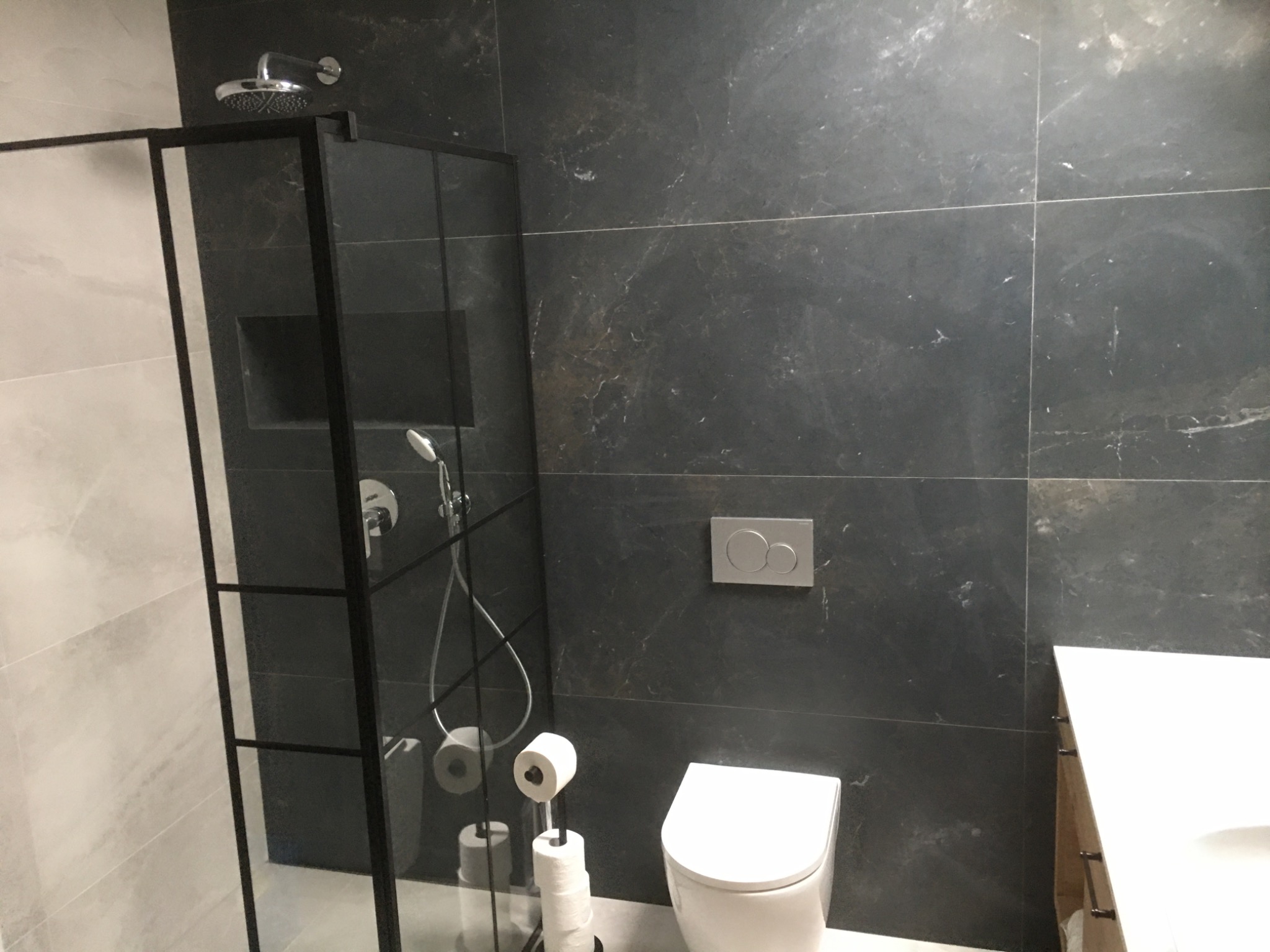 Nowoczesna łazienka z kabiną prysznicową typu walk-in z czarnymi profilami, toaletą wiszącą i umywalką nablatową na tle ciemnych, marmurkowych płytek.