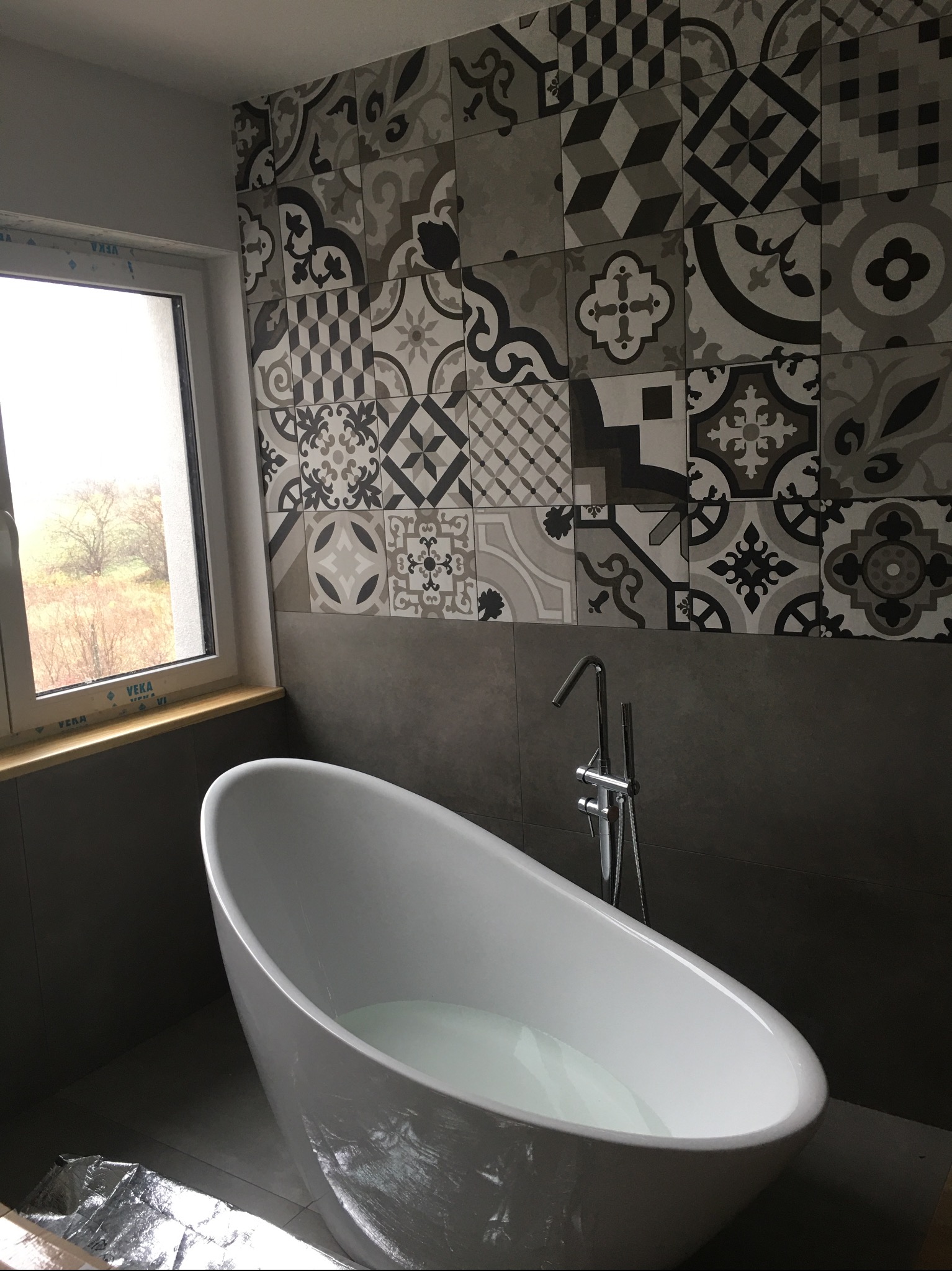 Nowoczesna łazienka z wolnostojącą wanną wypełnioną wodą, szare płytki z wzorami na ścianie, okno z widokiem na drzewa, chromowana bateria wannowa.
