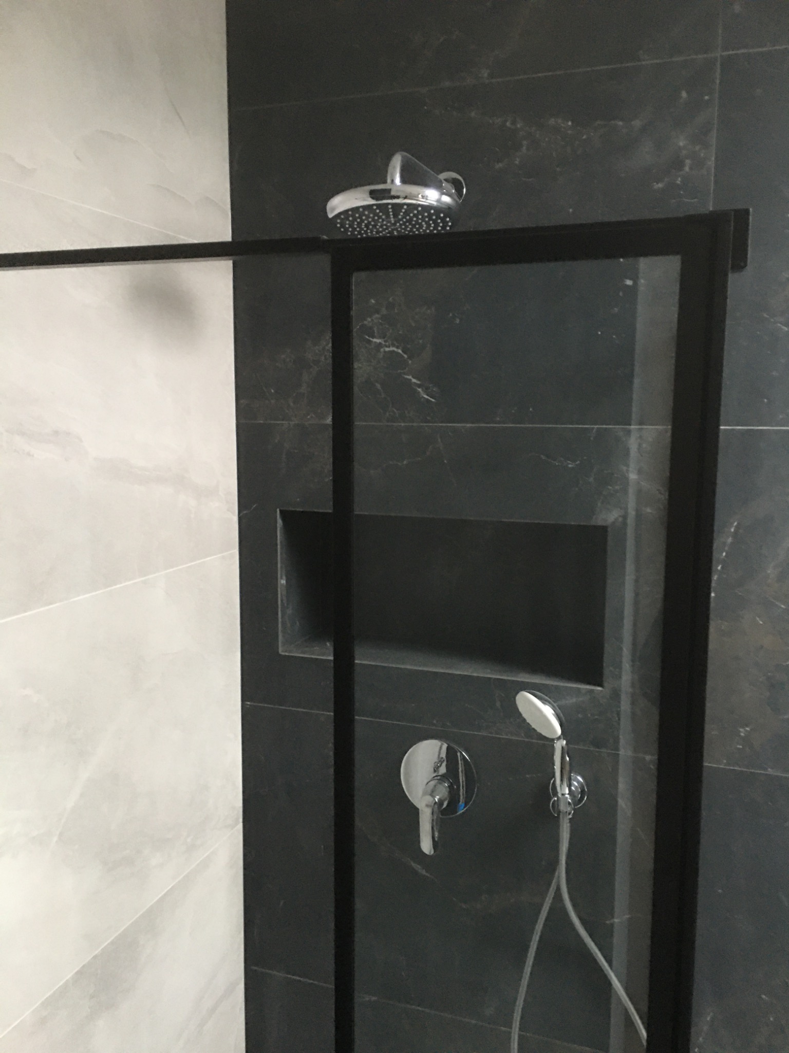 Nowoczesna kabina prysznicowa z czarną ramą, deszczownicą i panelem prysznicowym na tle szarych płytek imitujących marmur, z wnęką na kosmetyki.