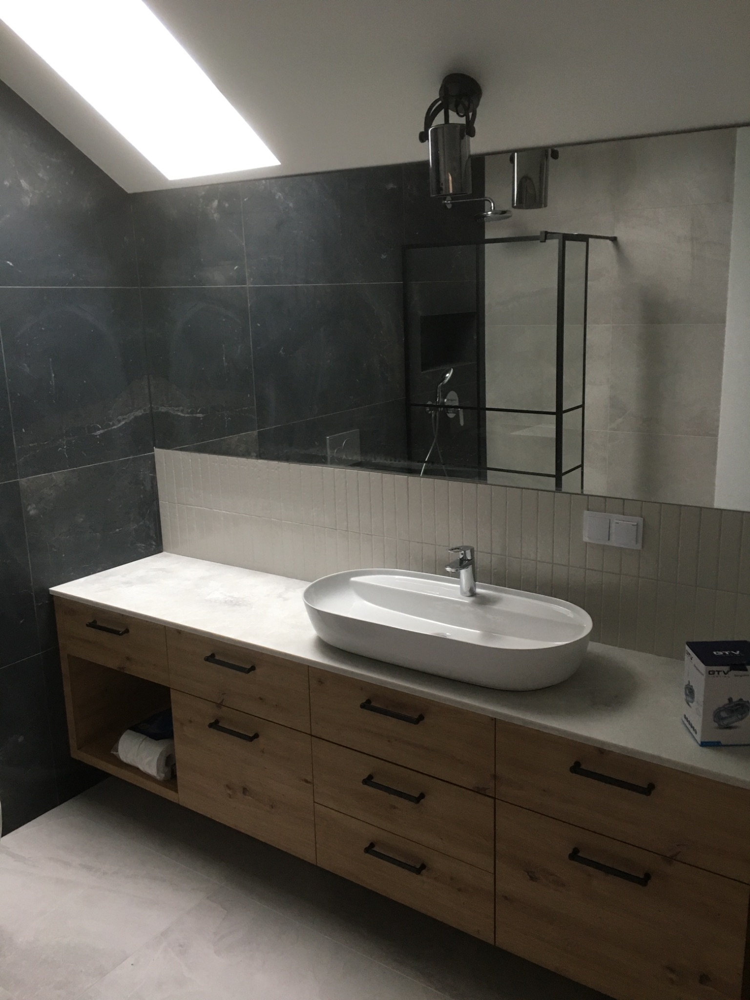 Nowoczesna łazienka w odcieniach szarości i drewna: owalna umywalka nablatowa, szafka z drewnianymi frontami, czarne płytki na ścianie i kabina prysznicowa z czarnymi ramami.