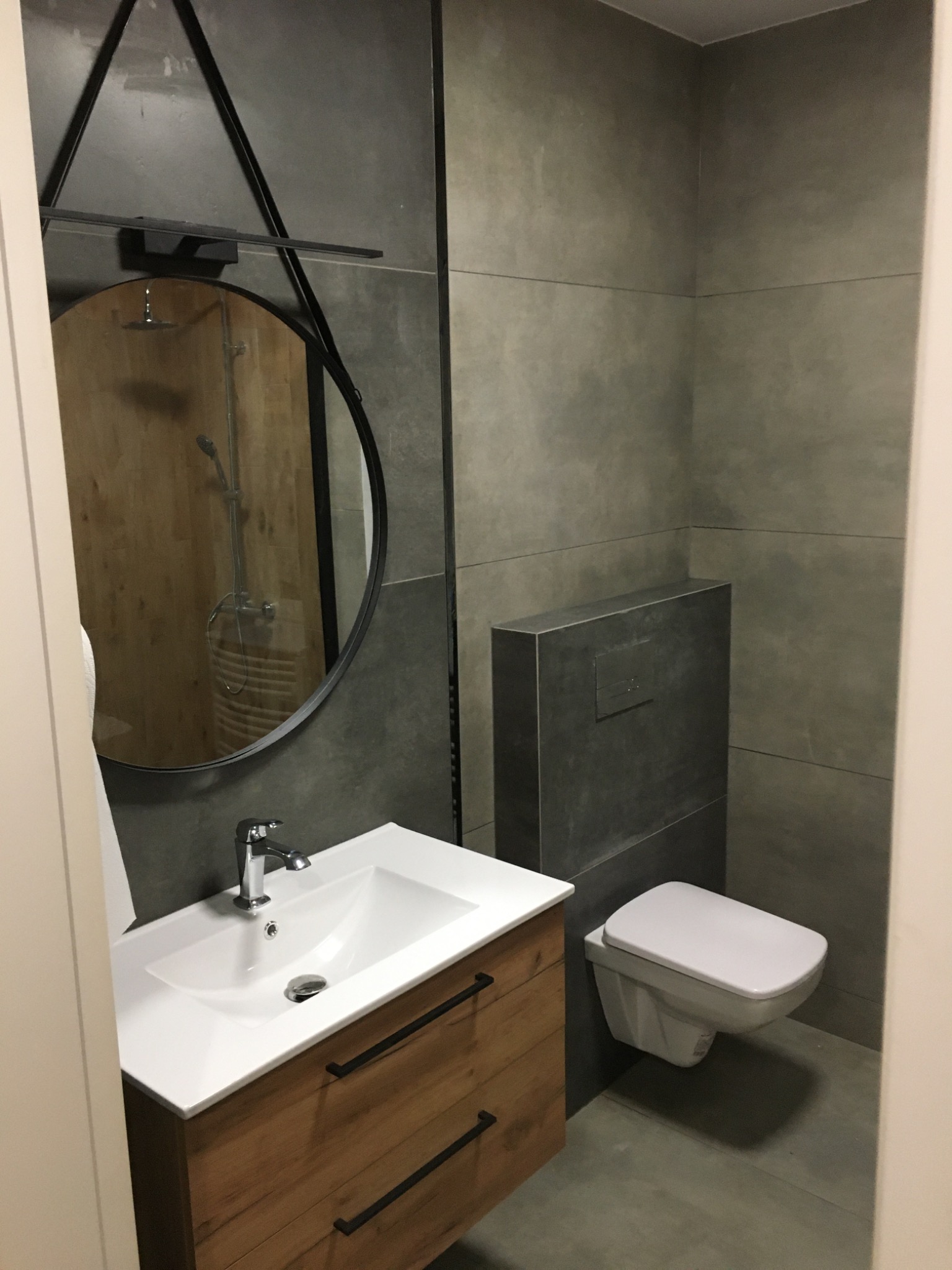 Nowoczesna łazienka z szarymi płytkami, wiszącą toaletą i umywalką na drewnianej szafce; lustro okrągłe z czarną ramą odbija prysznic.