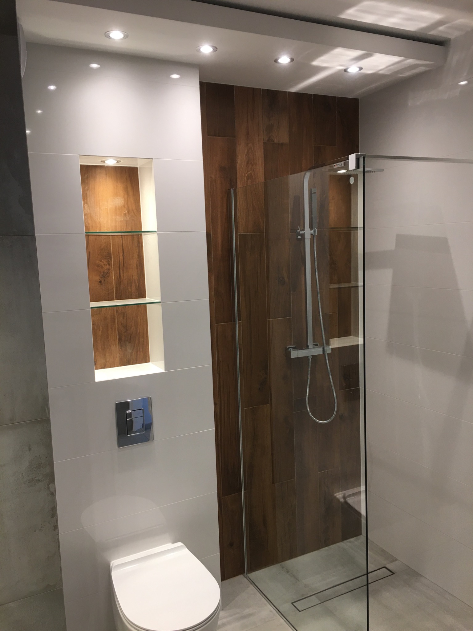 Nowoczesna łazienka z kabiną prysznicową typu walk-in, drewnopodobną ścianą z wnęką z półkami, białą ceramiką i oświetleniem punktowym.