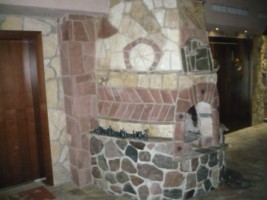 Kominek obłożony kamieniem naturalnym w odcieniach beżu, brązu i szarości, z widoczną strukturą i różnorodnością kształtów kamieni. W tle drzwi drewniane i fragment pomieszczenia z oświetleniem...