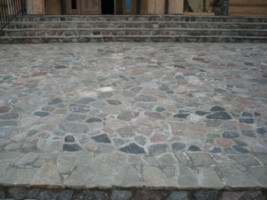 Nieregularna nawierzchnia z kamienia naturalnego w odcieniach szarości i różu, prowadząca do schodów wejściowych z kamiennymi stopniami i drewnianymi drzwiami w tle.