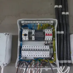 Otwarta skrzynka rozdzielcza z widocznymi bezpiecznikami, wyłącznikami, przewodami o różnych kolorach i grubościach, oraz sztywnymi rurami osłonowymi przymocowanymi do ściany. Obok widoczna...