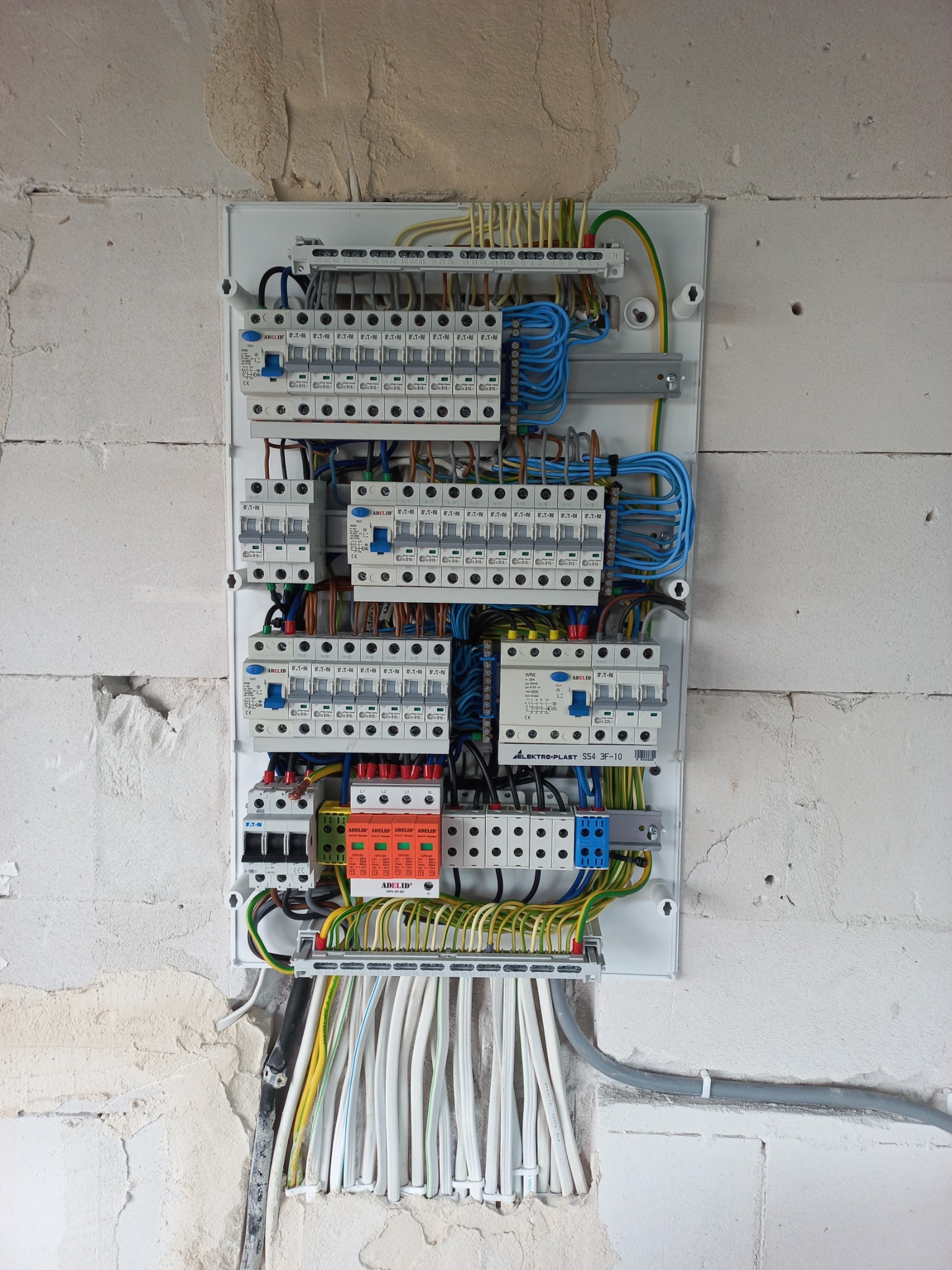 Rozdzielnica elektryczna z zainstalowanymi bezpiecznikami i okablowaniem, widoczna na tle surowej ściany z betonu komórkowego.