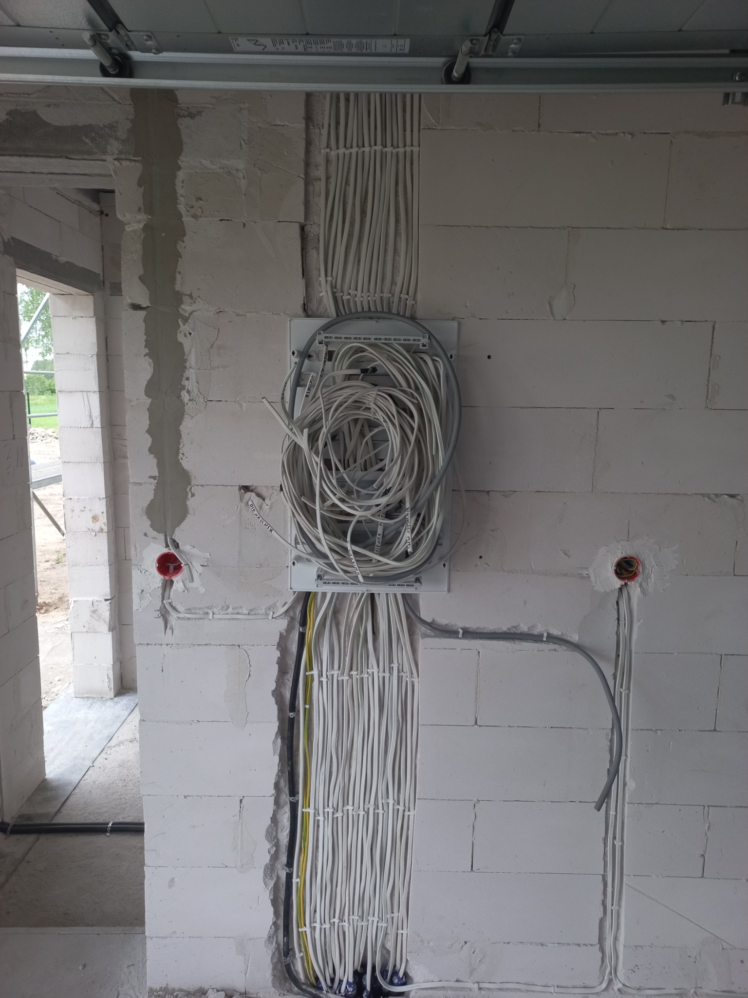 Rozdzielnica elektryczna z pętlą kabli w surowym wnętrzu budynku z białego bloczka, widoczne kable w peszlach oraz puszki instalacyjne.