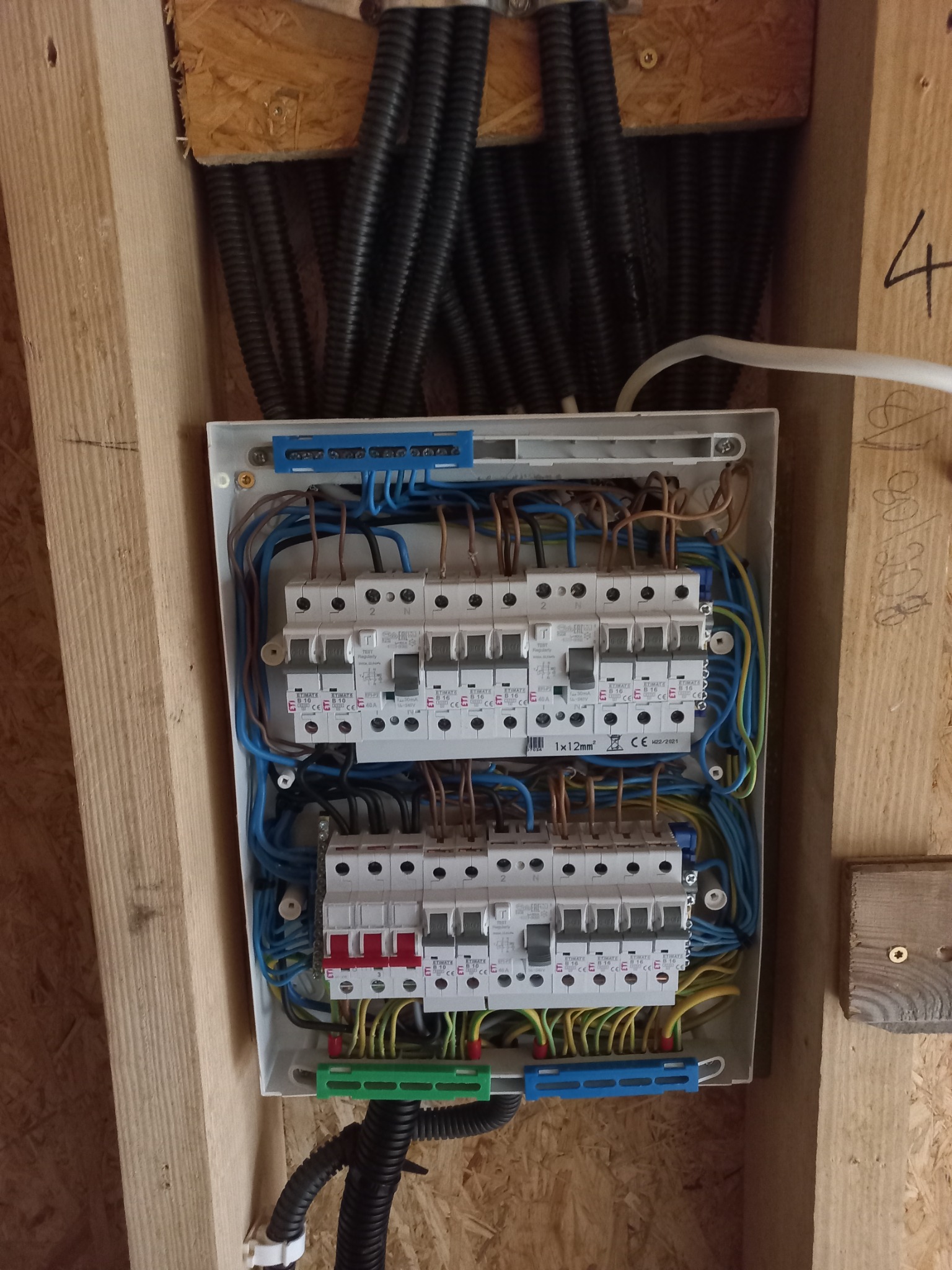 Otwarta skrzynka bezpiecznikowa z zainstalowanymi bezpiecznikami i przewodami, zamontowana na drewnianej ścianie z widocznymi rurami karbowanymi w tle.