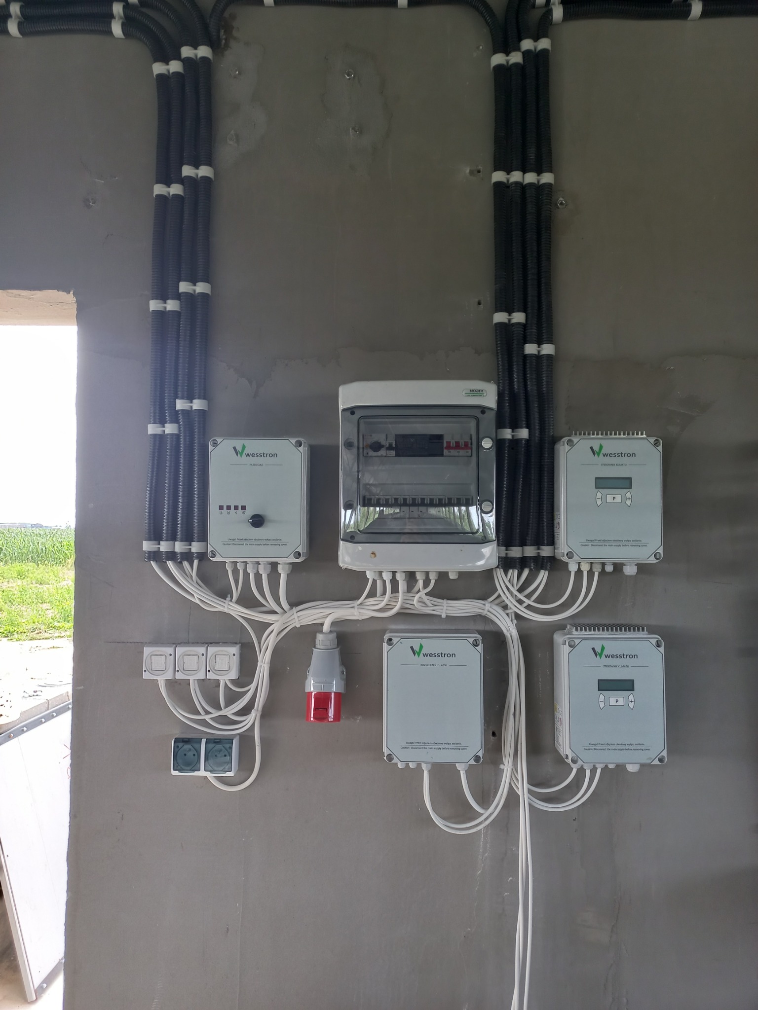 Szara ściana z zamontowaną rozdzielnicą elektryczną, czterema modułami pomiarowymi firmy Wesstron, gniazdami oraz peszlami z przewodami. Widok na zewnątrz przez otwarty otwór w ścianie.