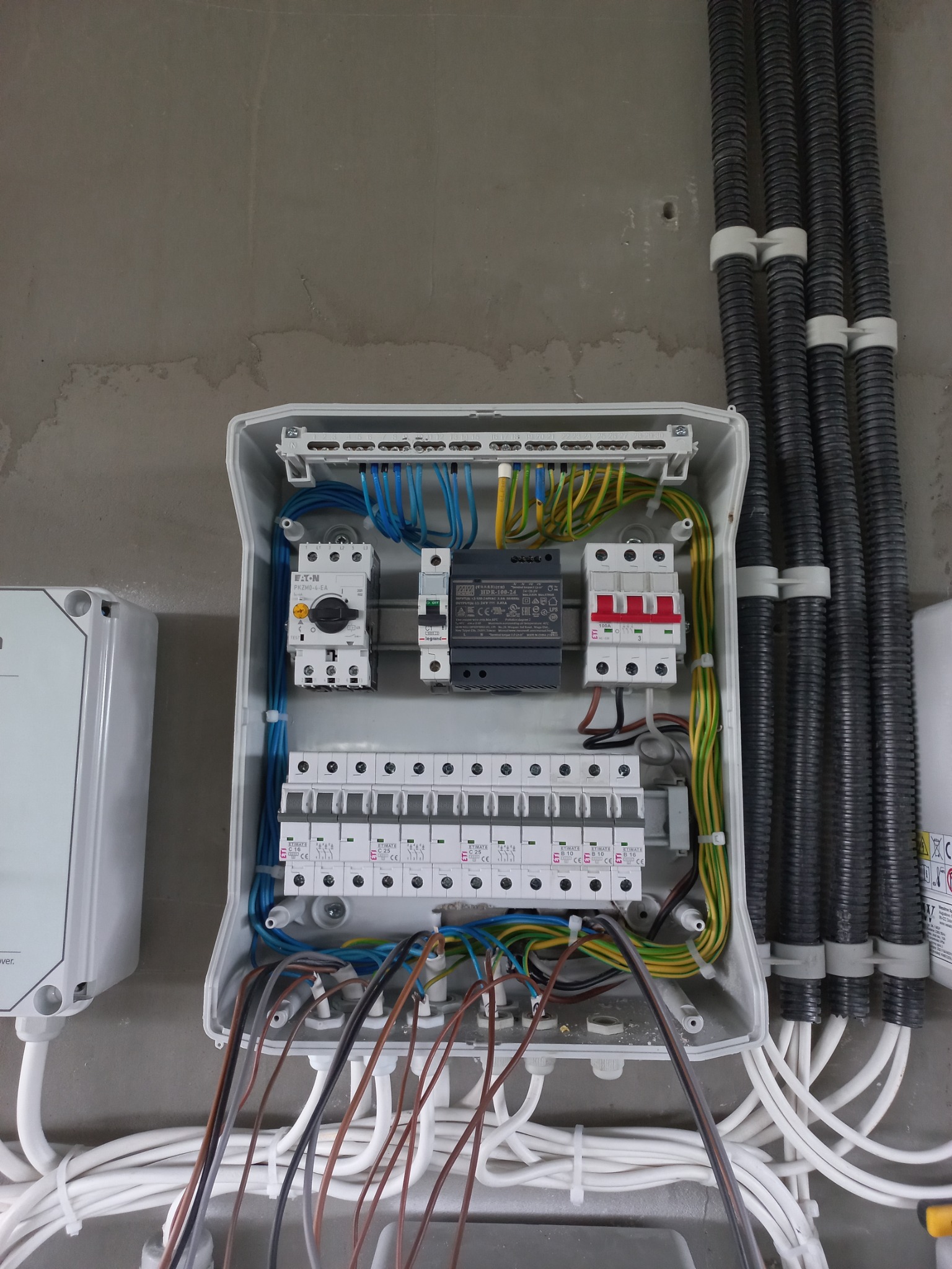 Otwarta skrzynka rozdzielcza z widocznymi bezpiecznikami, wyłącznikami, przewodami o różnych kolorach i grubościach, oraz sztywnymi rurami osłonowymi przymocowanymi do ściany. Obok widoczna...