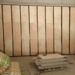 Ściana z zamontowanymi panelami izolacyjnymi w metalowej ramie, widoczne paczki materiałów budowlanych i stos płyt na palecie, surowy stan wykończenia wnętrza.