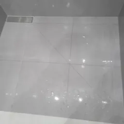 Wykończona, jasnoszara kabina prysznicowa z odpływem liniowym, widoczne refleksy światła na płytkach podłogowych i ściennych.