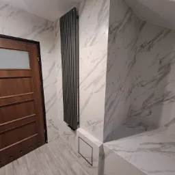 Wykończona łazienka z marmurowymi płytkami, grzejnikiem panelowym w kolorze antracytowym i drewnianymi drzwiami z szybą.