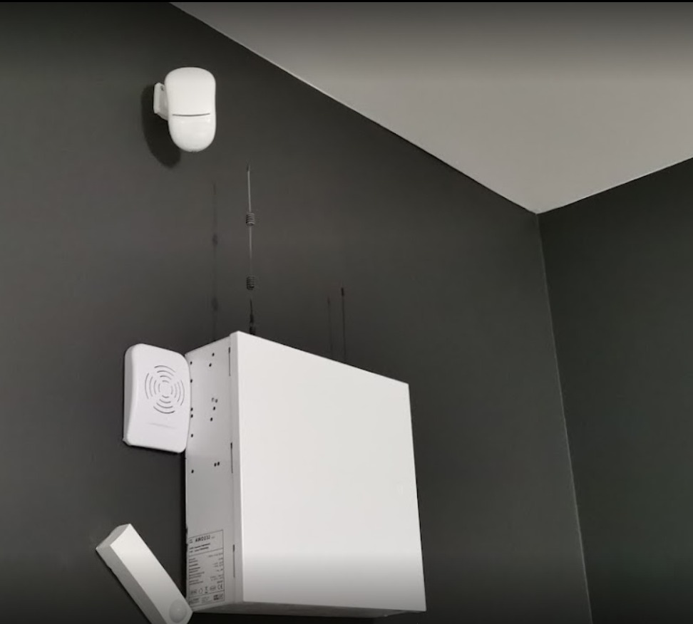 Centrala alarmowa, czujnik ruchu i sygnalizator dźwiękowy zamontowane na ciemnoszarej ścianie w rogu pomieszczenia.
