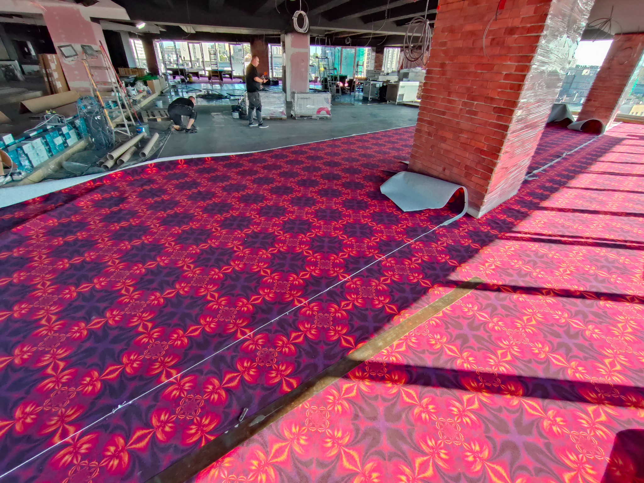 Układanie wzorzystej, czerwono-fioletowej wykładziny w przestronnym wnętrzu z ceglanymi kolumnami. Widoczni pracownicy podczas pracy, narzędzia i materiały budowlane.