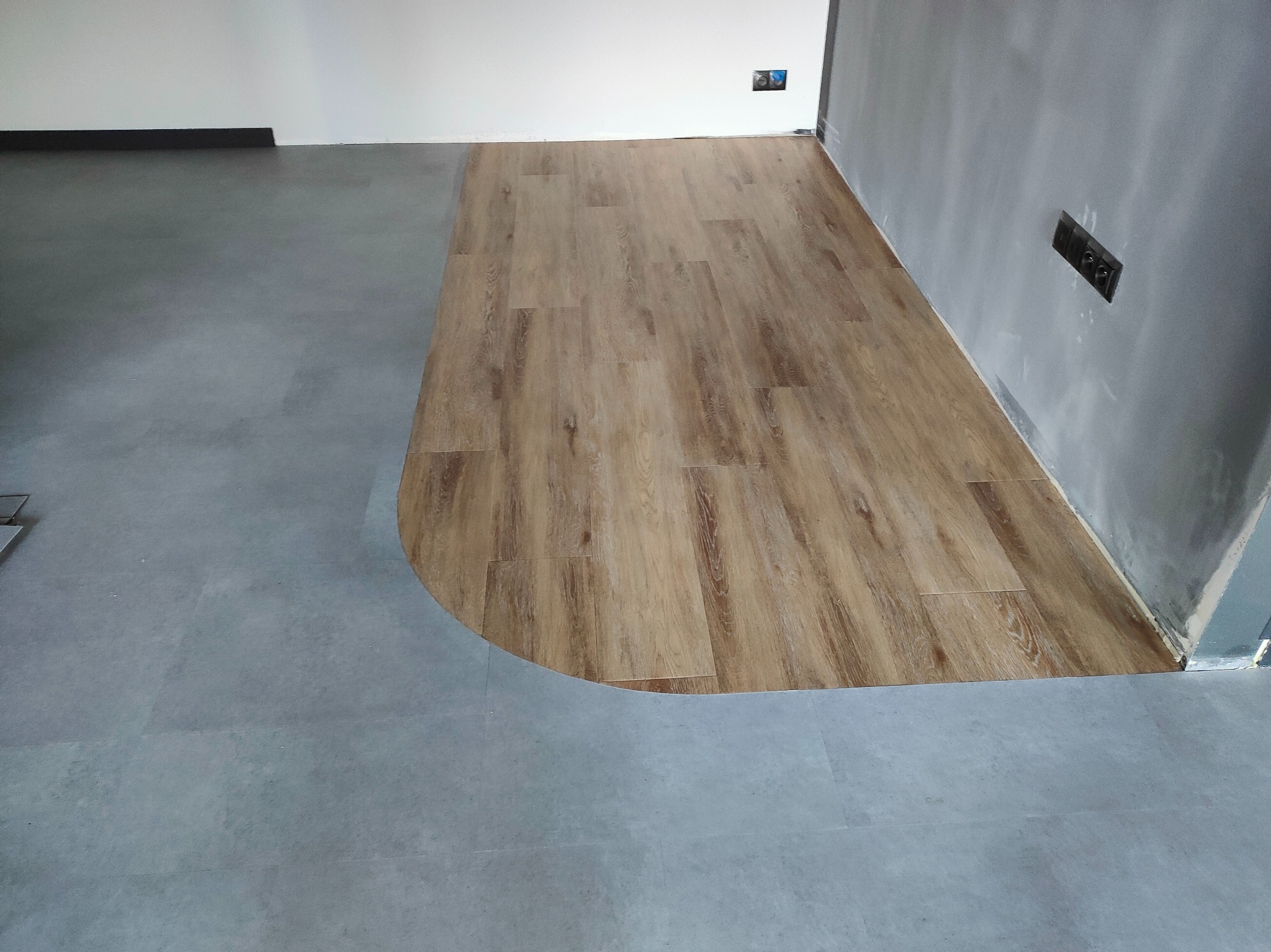 Połączenie szarej wykładziny i paneli drewnopodobnych w rogu pomieszczenia. Widoczne gniazdka elektryczne na szarej ścianie. Podłoga po remoncie.