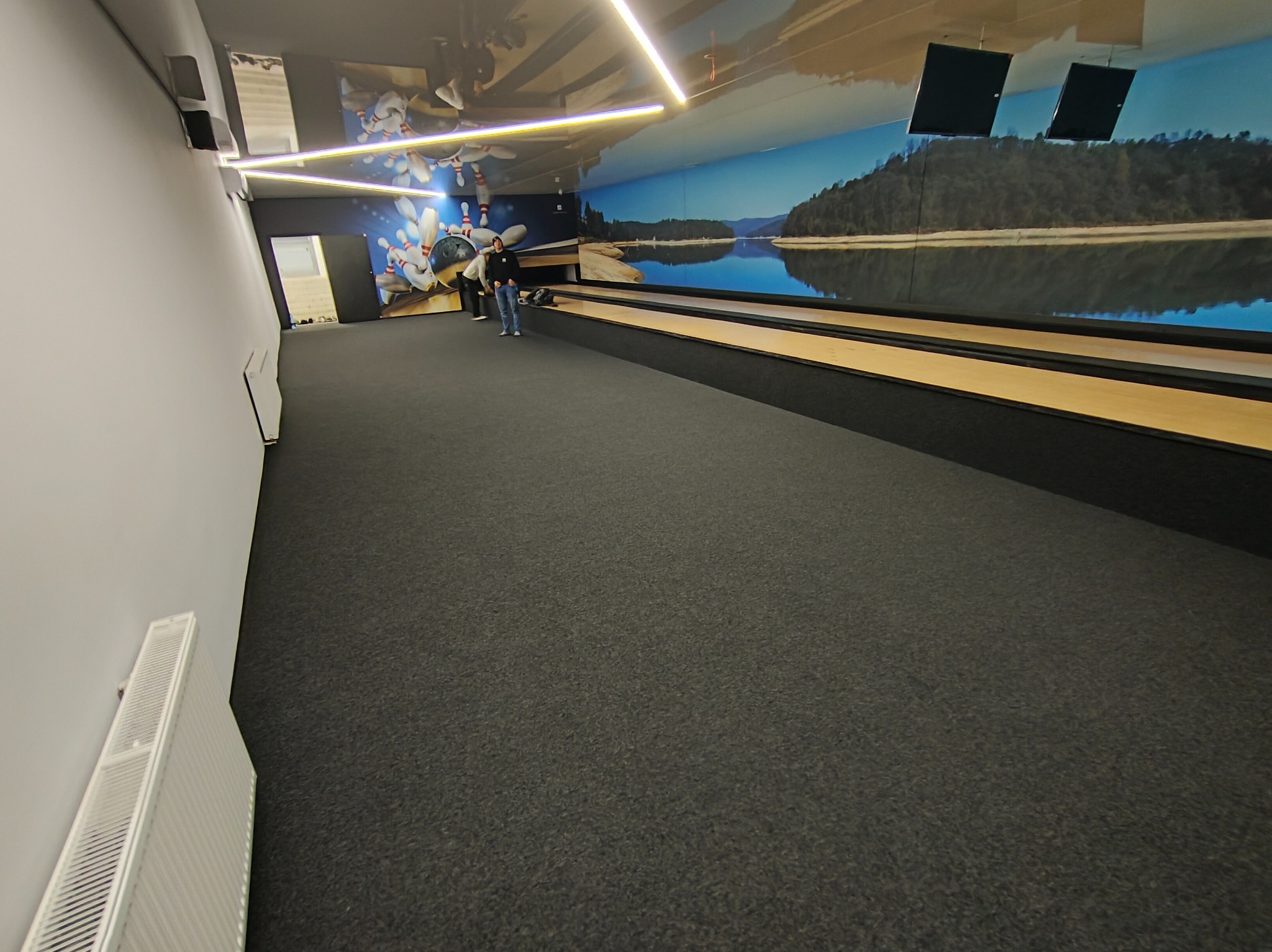 Ciemnoszara wykładzina dywanowa w kręgielni z motywem jeziora na ścianie. Widoczne tory do gry i osoby w tle. Oświetlenie LED na suficie.