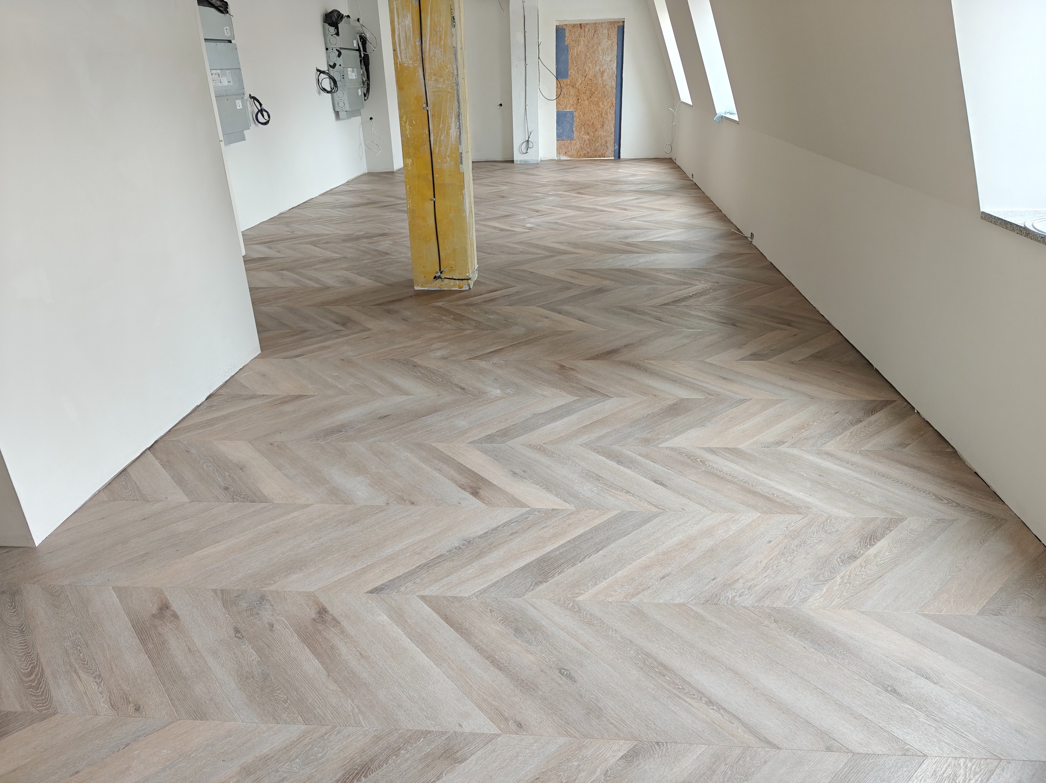 Jasne panele ułożone w jodełkę francuską w przestronnym, jasnym pokoju na poddaszu. Widoczny fragment kolumny zabezpieczonej folią i kartonem.