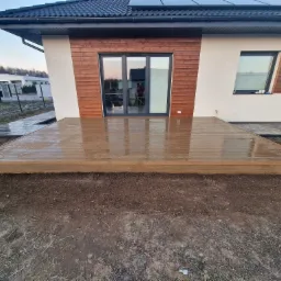 Nowoczesny, drewniany taras przydomowy z widocznymi refleksami wody po deszczu, z brązową elewacją i oknami domu w tle.