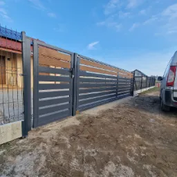 Nowoczesne ogrodzenie panelowe z drewnianymi akcentami, brama otwarta. Widoczny dom z panelami słonecznymi i auto dostawcze. Słoneczny dzień.