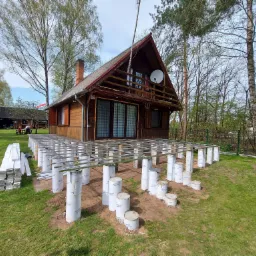 Przygotowanie podłoża pod taras przy drewnianym domu: betonowe słupy fundamentowe z metalową konstrukcją nośną, deski tarasowe obok, widok na dom z anteną satelitarną.