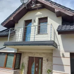 Elewacja domu z balkonem z metalową balustradą, brązowe okna, drzwi wejściowe z kamienną okładziną, panele słoneczne na dachu.