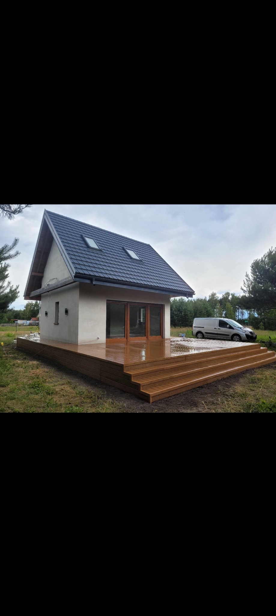 Nowoczesny dom z beżową elewacją i dużymi przesuwnymi drzwiami, otoczony tarasem z brązowych desek kompozytowych z kilkoma stopniami. Na podjeździe zaparkowany biały samochód dostawczy.