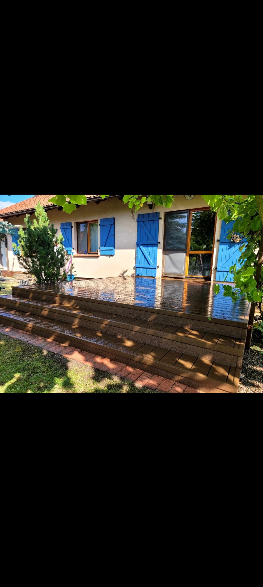 Nowoczesny taras z desek kompozytowych w odcieniach brązu, przylegający do domu z błękitnymi okiennicami i ceglaną obwódką, lekko mokry od deszczu.