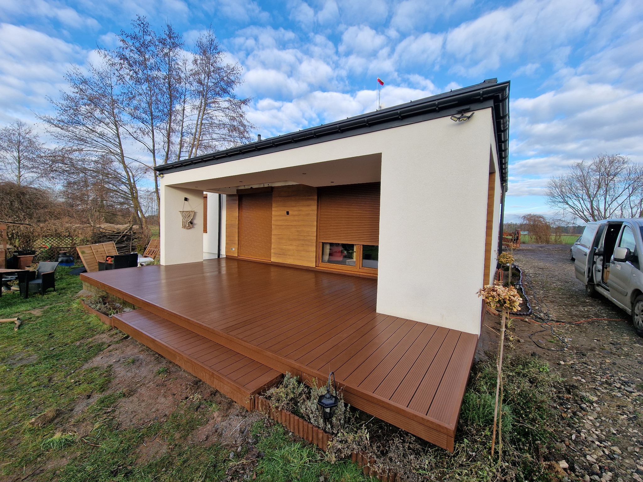 Nowoczesny taras z brązowych desek kompozytowych z dwustopniowymi schodami, przylegający do białego budynku z drewnianymi roletami, widok na pochmurne niebo i częściowo odsłonięty samochód dostawczy.