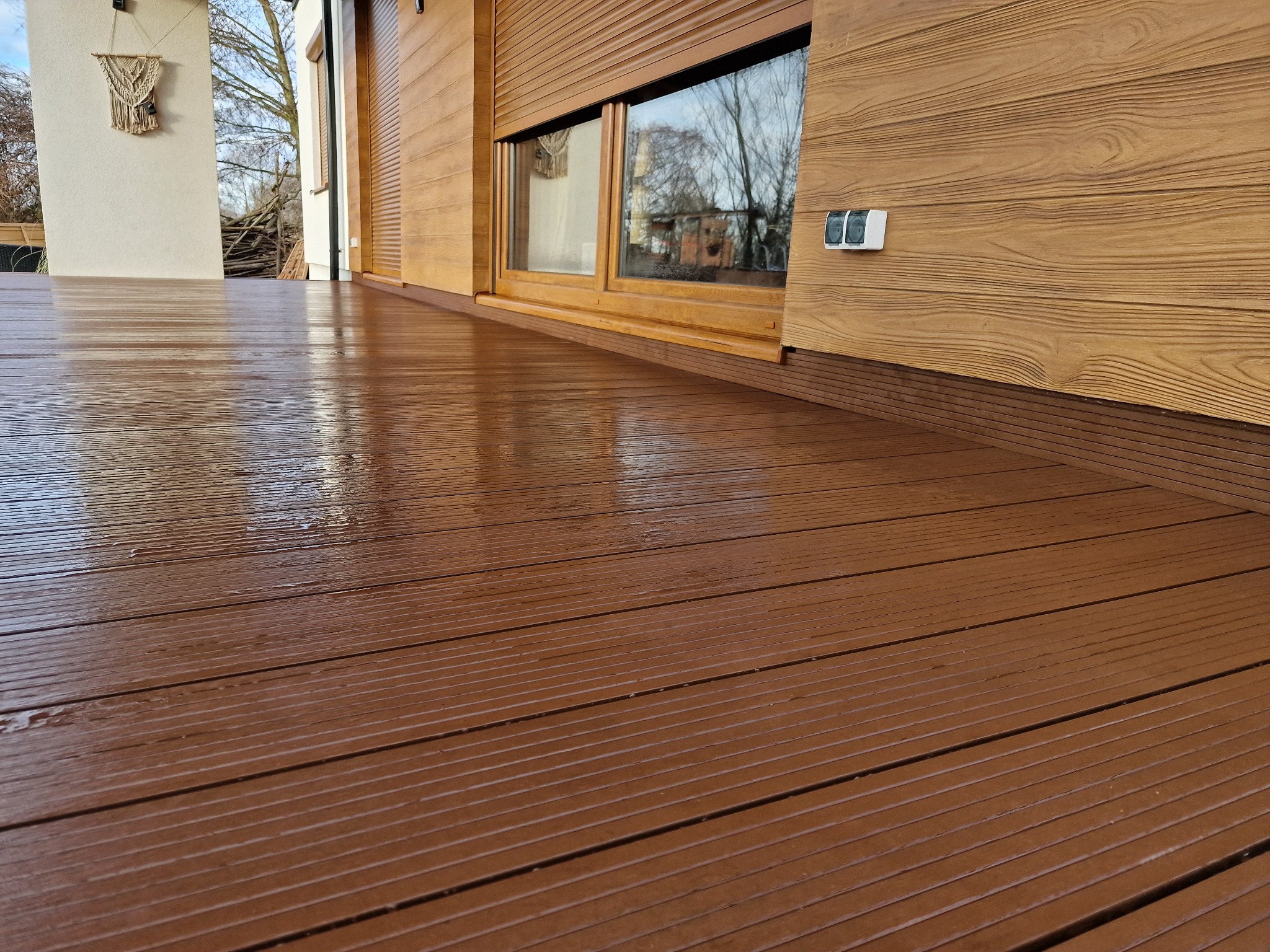 Brązowy taras kompozytowy z widoczną fakturą drewna, przylegający do ściany budynku z drewnianą elewacją i oknem z roletami zewnętrznymi.