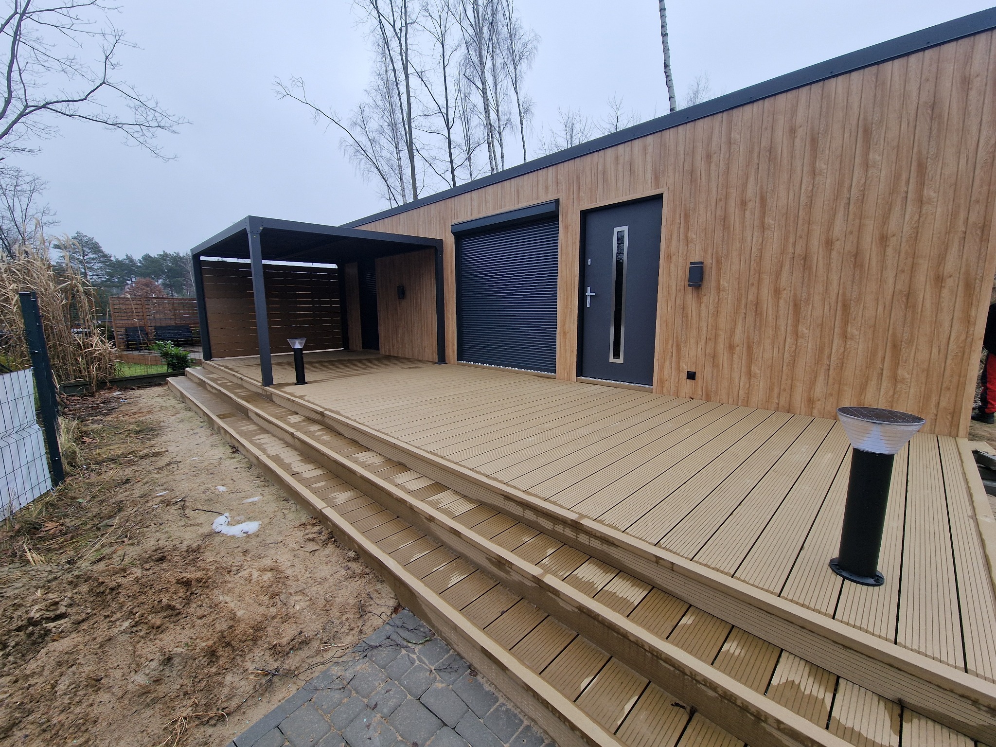 Nowoczesny dom z elewacją drewnopodobną, tarasem z desek kompozytowych i pergolą w kolorze antracytowym, oświetlony lampami solarnymi. Częściowo zrealizowany teren wokół tarasu.