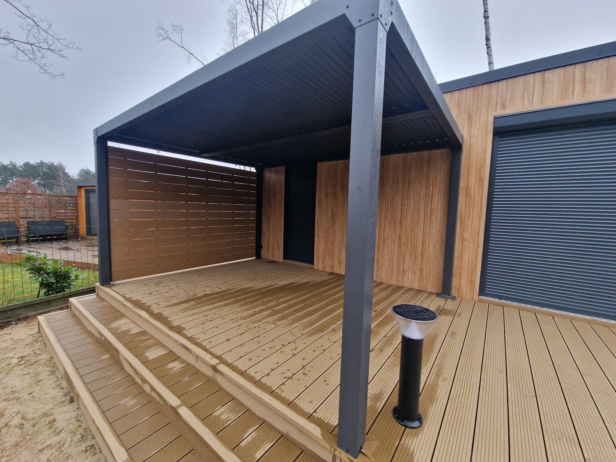 Zadaszona przestrzeń z tarasem kompozytowym i metalową pergolą, zintegrowana z elewacją budynku, oświetlona lampą solarną, widok na schody wejściowe i ogrodzenie w tle.