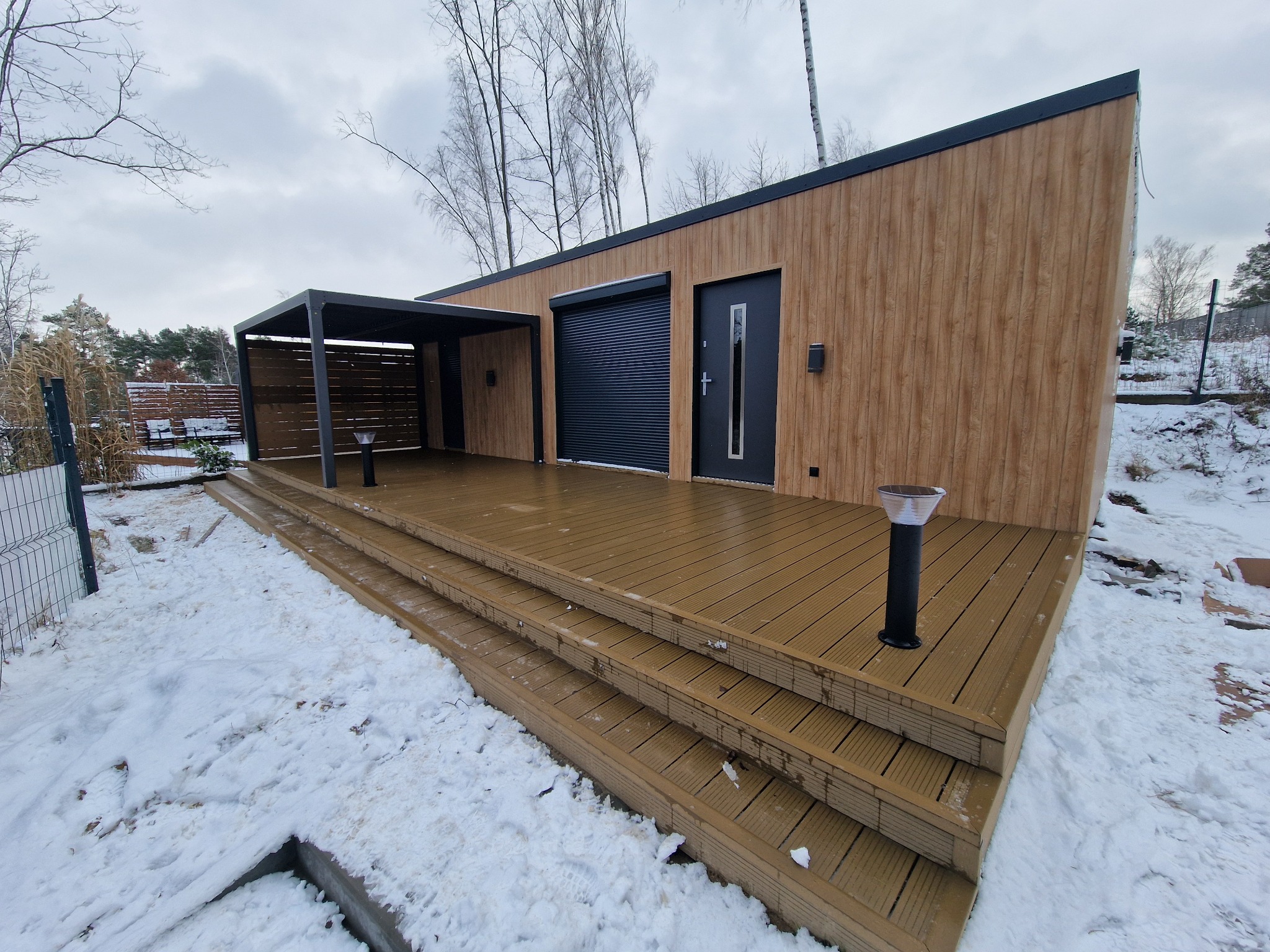 Nowoczesny budynek z elewacją drewnopodobną, zadaszeniem i tarasem z desek kompozytowych pokrytym śniegiem. Widoczne lampy solarne i zimowy krajobraz w tle.