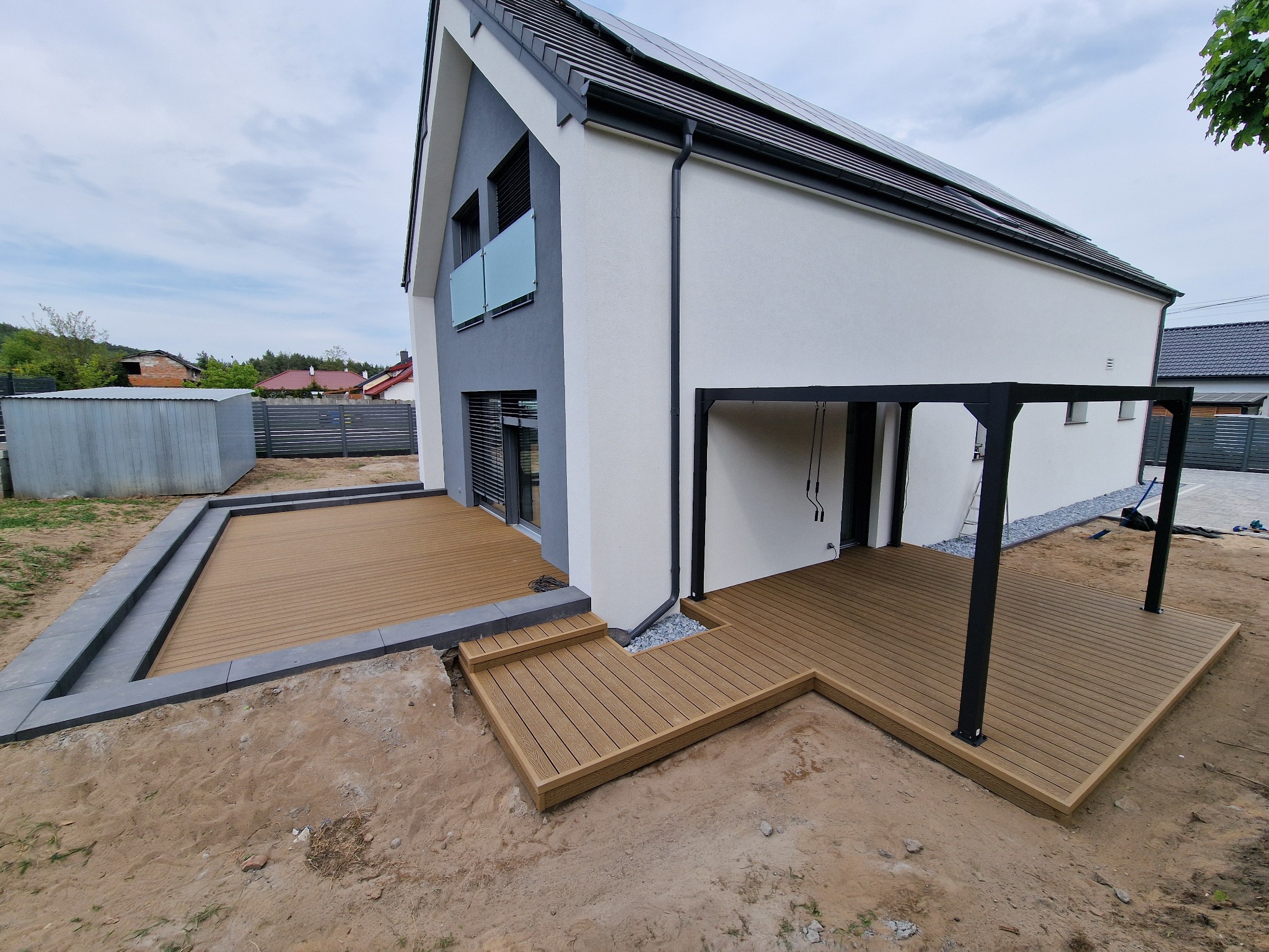 Nowoczesny taras kompozytowy przy białym domu z ciemną pergolą. Wykończone obrzeża, schodki prowadzące do domu. Widok z boku, ziemia wokół.