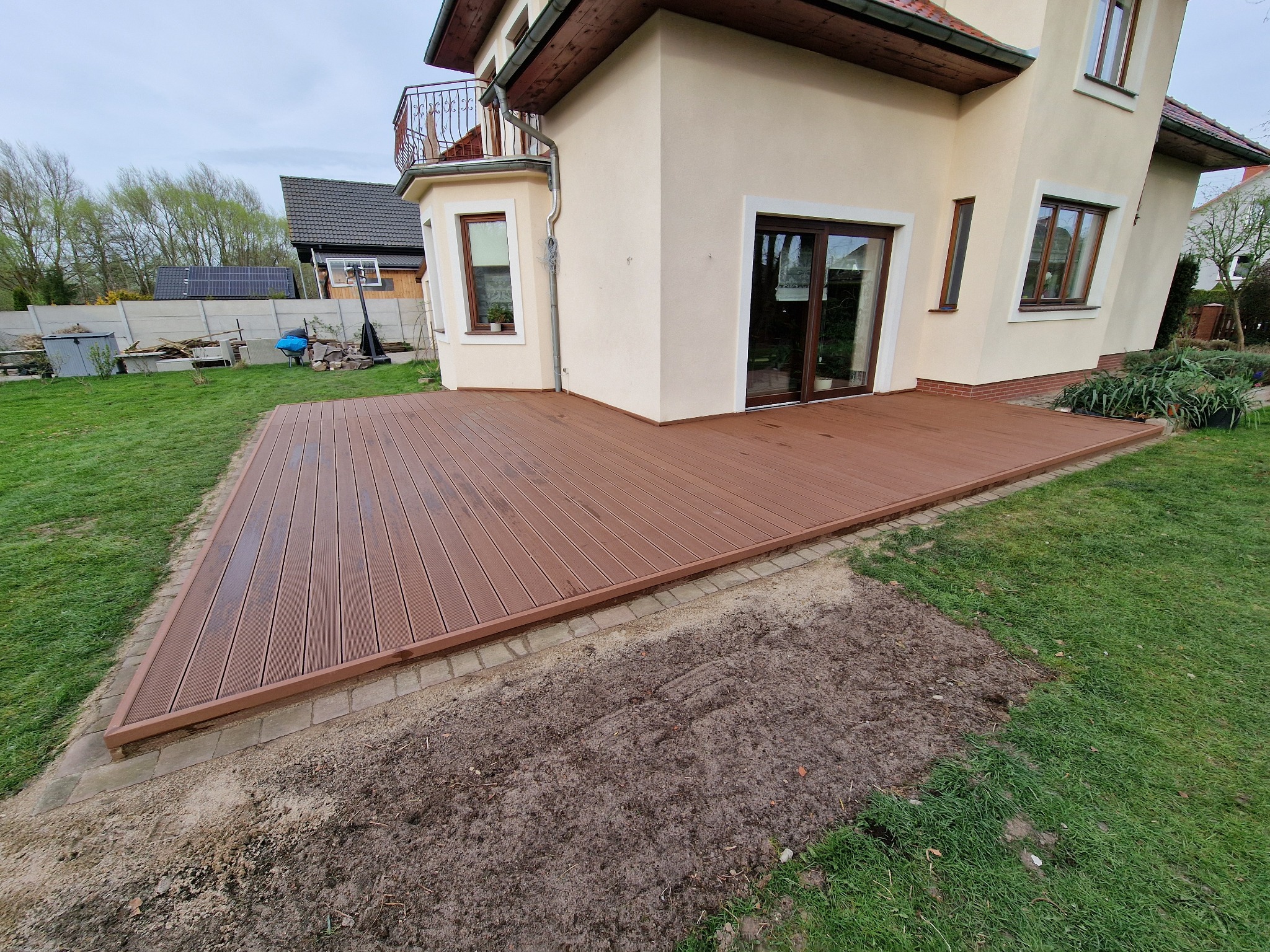 Brązowy taras kompozytowy przylegający do beżowego domu z balkonem. Widok na zielony trawnik i fragment ogrodu. Część obrzeża tarasu jeszcze nie wykończona.