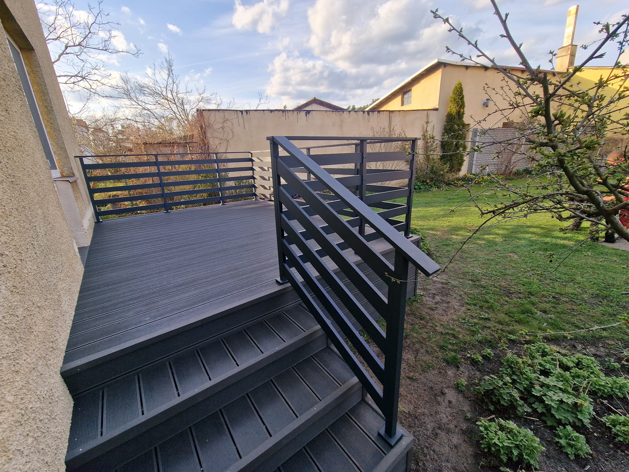 Nowoczesny taras z kompozytu w kolorze grafitowym, schody z barierką, widok na ogród i budynek w tle, słoneczny dzień.