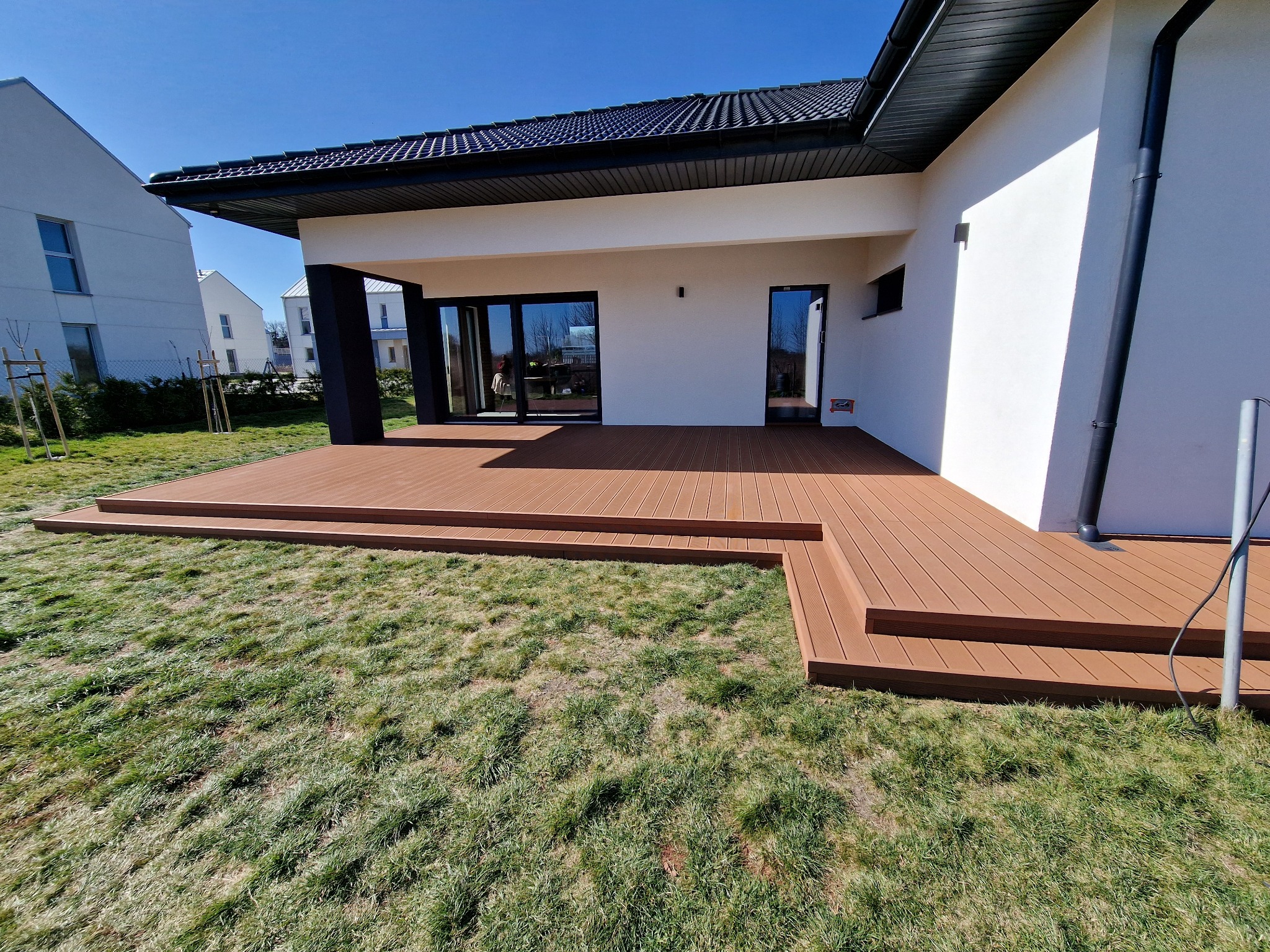 Nowoczesny taras kompozytowy z brązowych desek, z niskimi schodkami, przylegający do białego domu z czarnymi ramami okiennymi, otoczony zielonym trawnikiem.